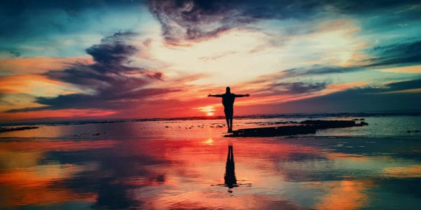 My Story Person standing with arms stretched on a beach at sunrise with red coloured sky reflected in the water.
